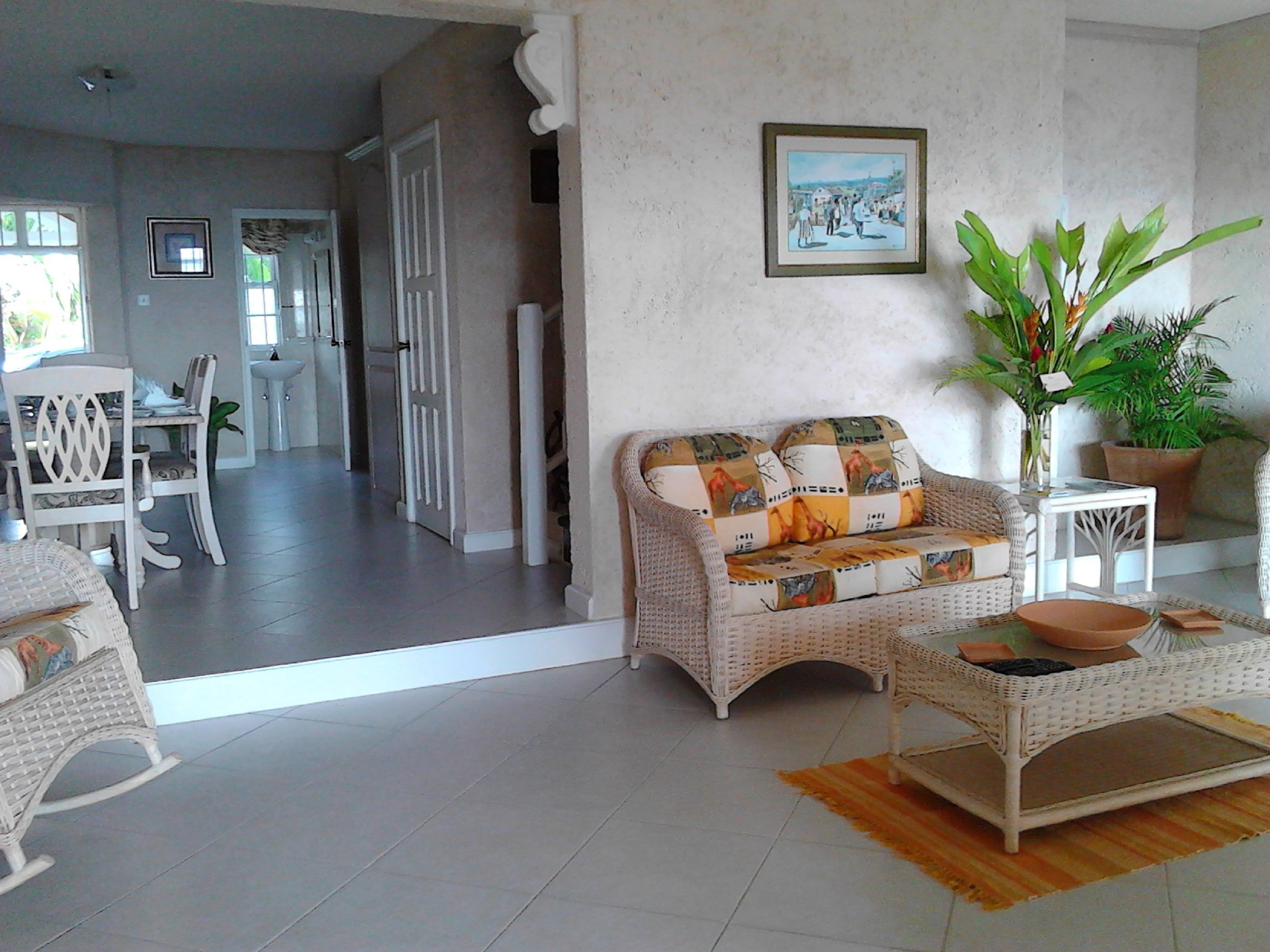 A living room with a wicker sofa, a dining table with chairs, a potted plant, and a framed picture on the wall.