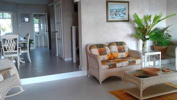 A living room with a wicker sofa, a dining table with chairs, a potted plant, and a framed picture on the wall.