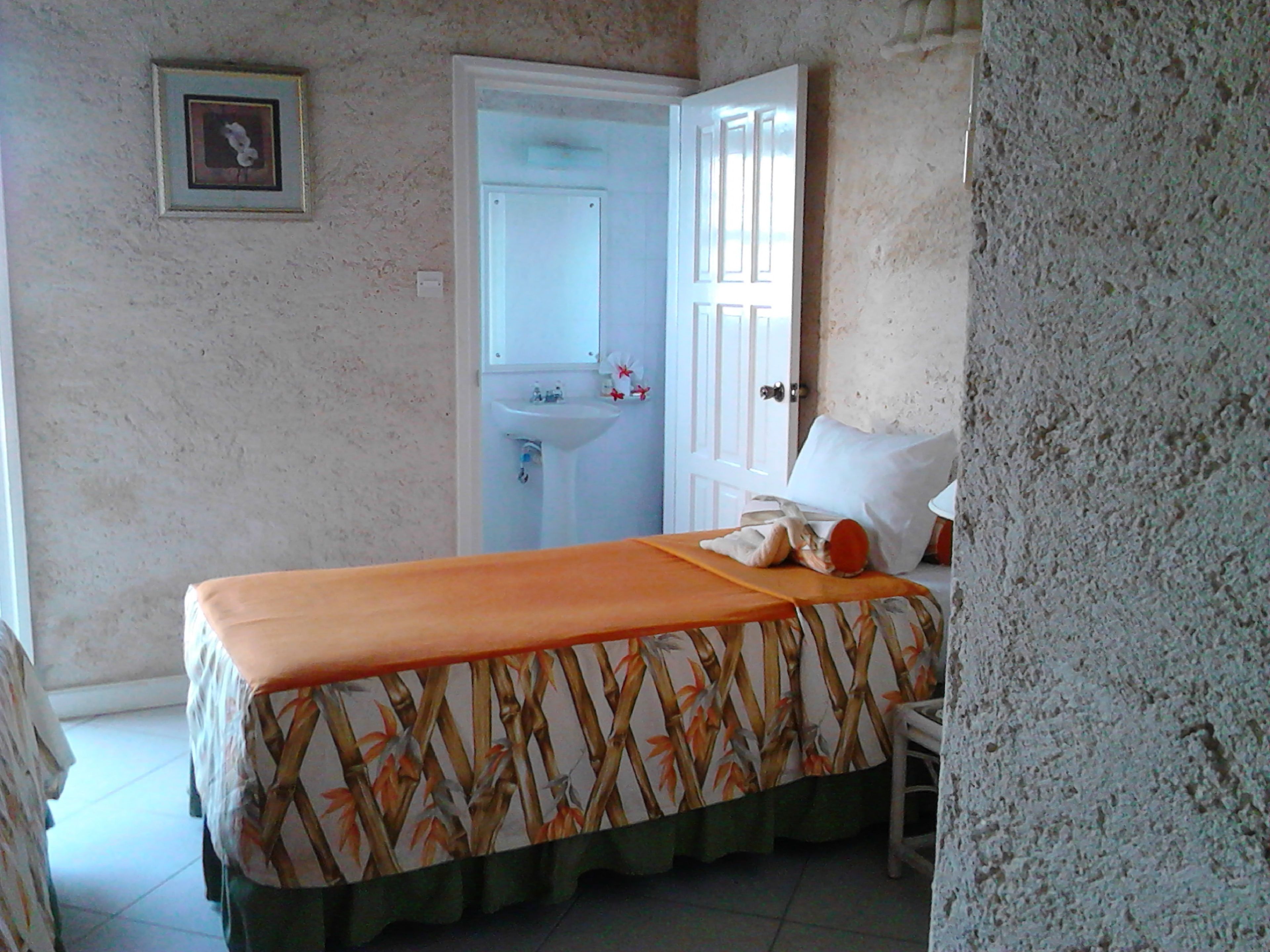 A bedroom with a bed, a nightstand, a bathroom with a sink and mirror, and a door leading to another room.
