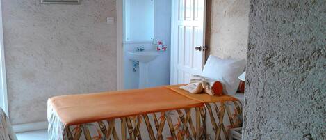 A bedroom with a bed, a nightstand, a bathroom with a sink and mirror, and a door leading to another room.