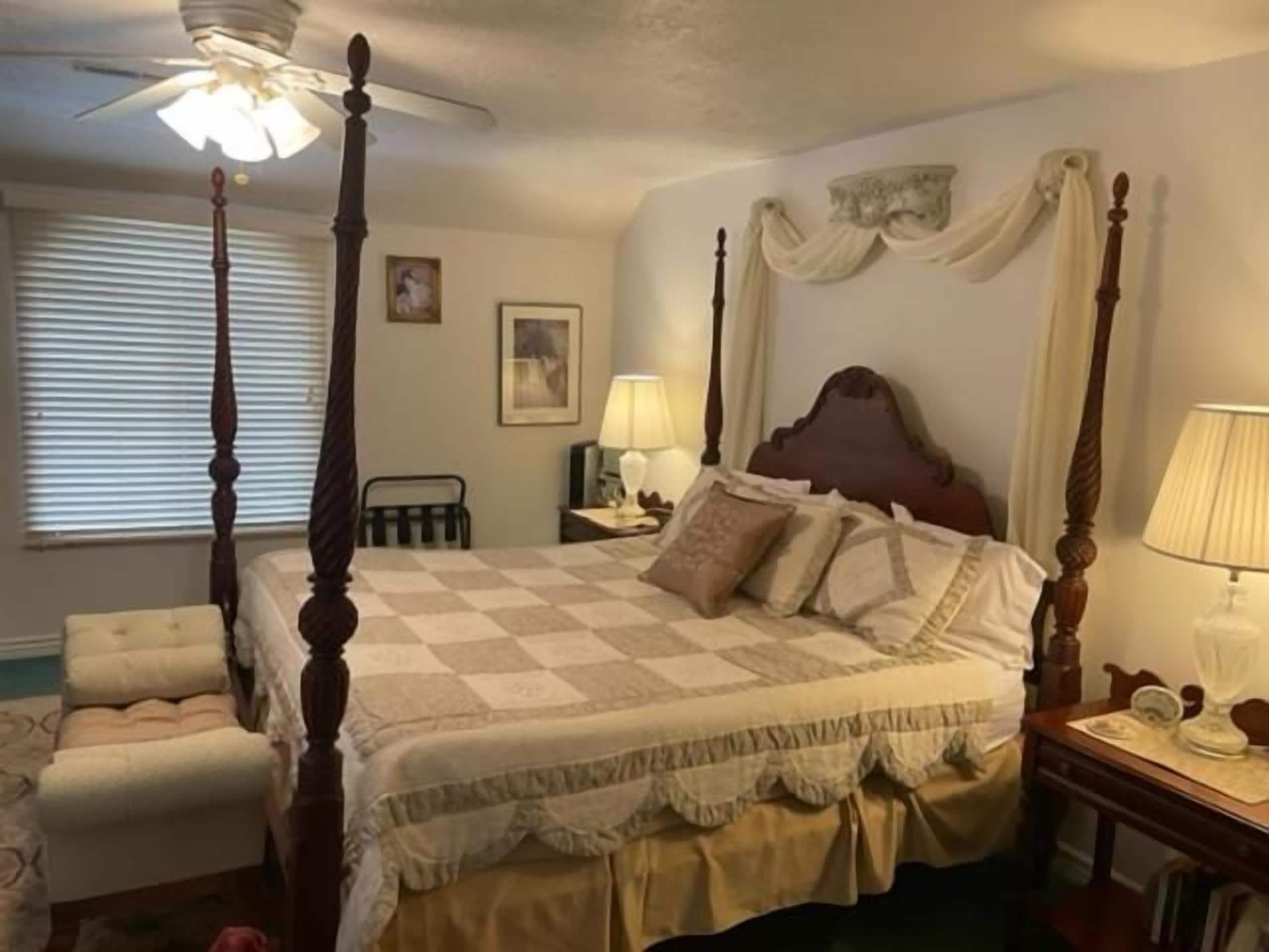 A four-poster bed with a checkered bedspread, a nightstand with a lamp, and a chair with a cushion.