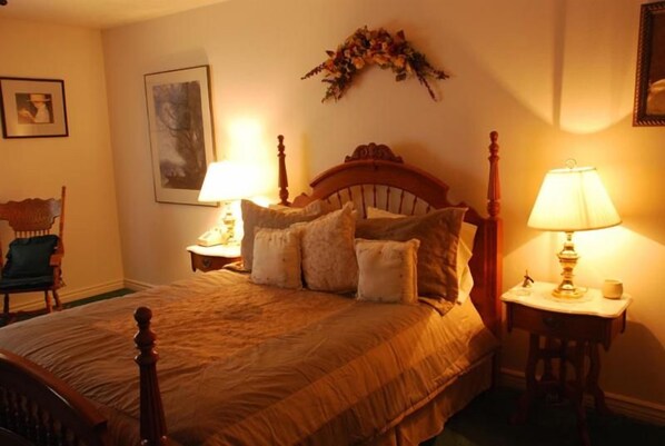 Quarto com cama de madeira, duas mesas de cabeceira com luminárias, uma cadeira e um objeto decorativo na parede.