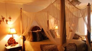 Un dormitorio con una cama con dosel, una mesita de noche con lámpara y una ventana con cortinas.