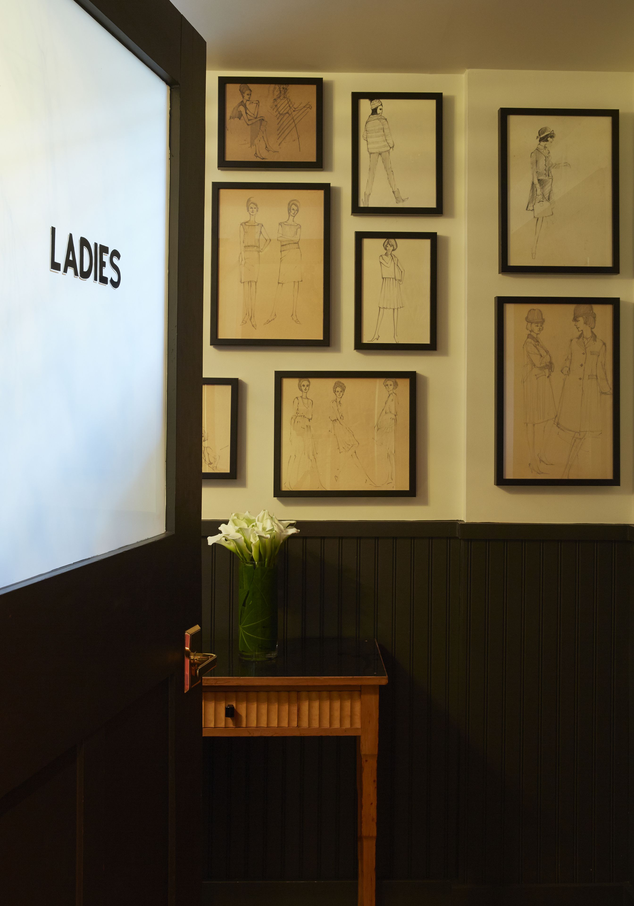 A wall with framed sketches of women in various poses, a wooden table with a vase of flowers, and a projection screen displaying the word "LADIES".