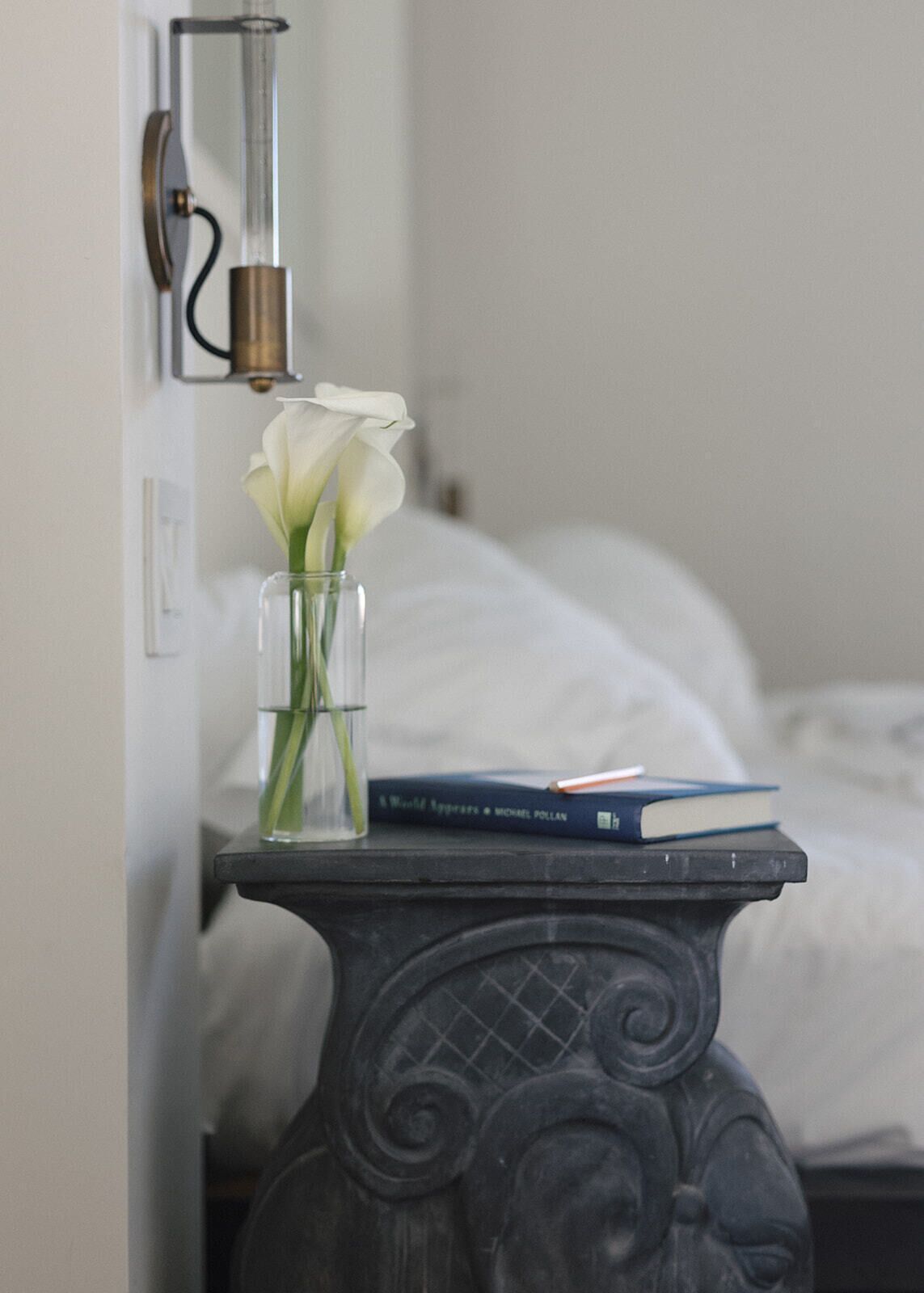 Una mesita de noche con un jarrón de flores blancas, un libro y una lámpara.