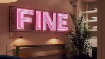 A room with a neon sign reading "FINE" on the wall, a desk with a lamp, a plant, and a bookshelf.