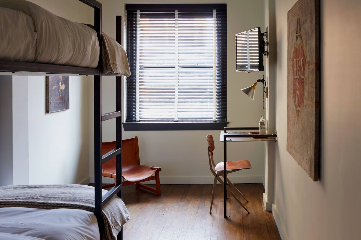 A dormitory room with bunk beds, a desk, a chair, and a window with blinds.