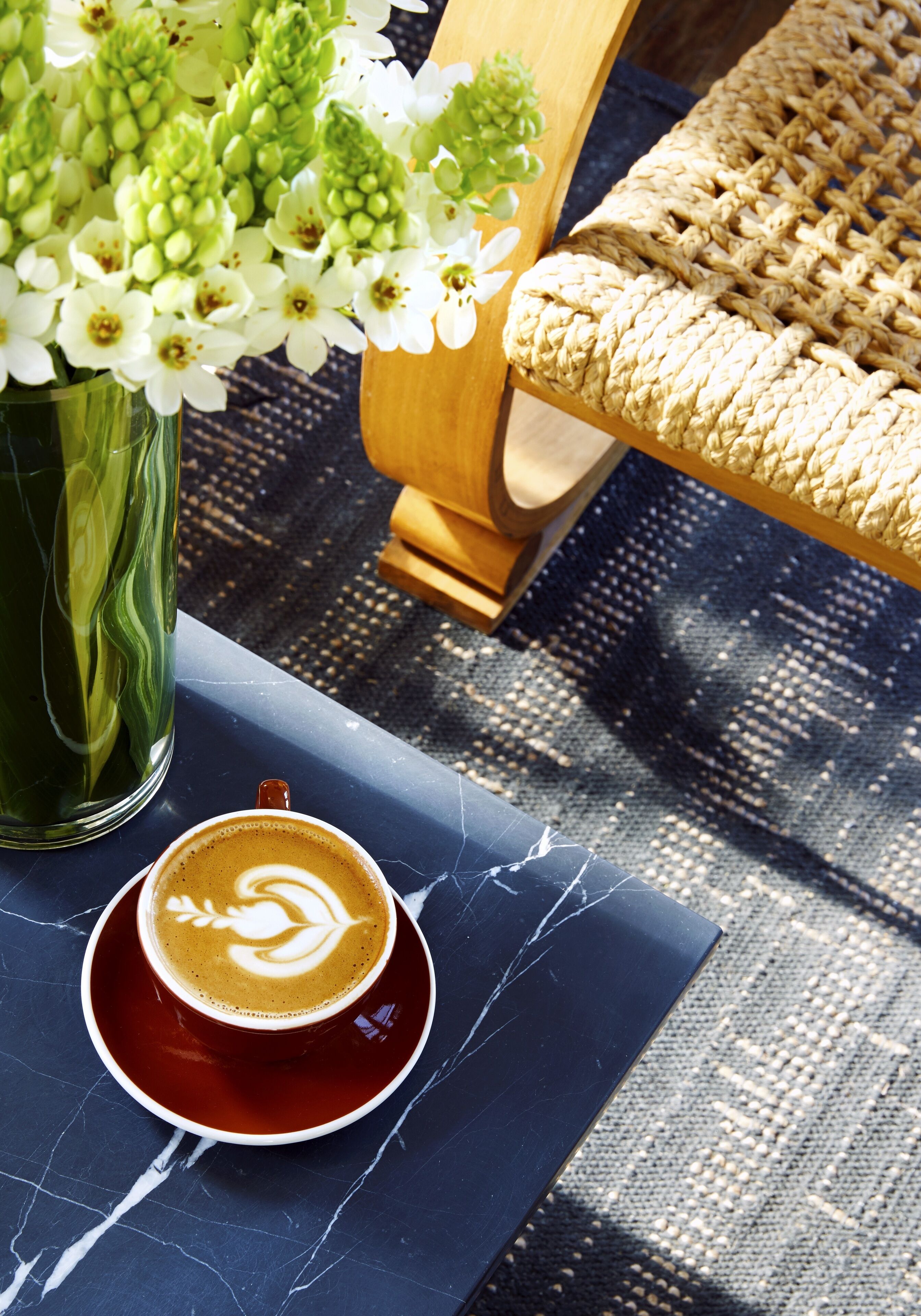 Una taza de café con arte de latte sobre una mesa de mármol oscuro, un jarrón verde con flores blancas y una silla de mimbre.