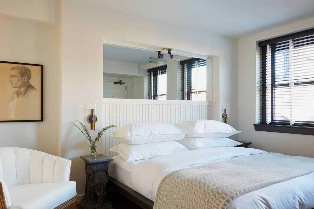 A bedroom with a large bed, a chair, a mirror, and a window with blinds.