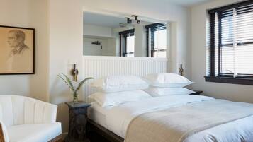 A bedroom with a large bed, a chair, a mirror, and a window with blinds.