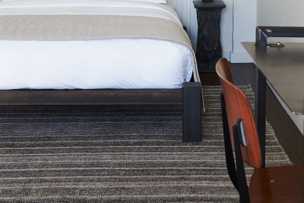 A modern hotel room with a large bed, a desk, a chair, a small table, a potted plant, and a mirror.
