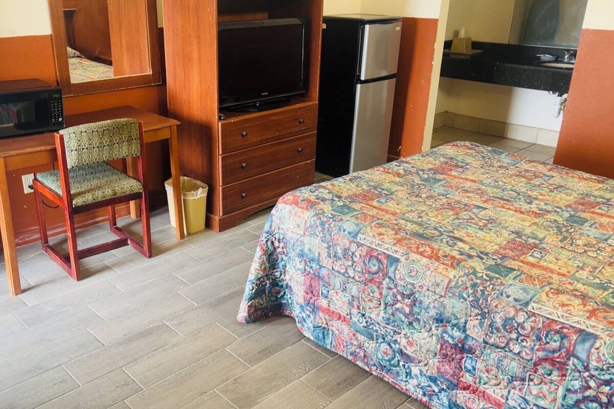 A hotel room with a bed, a desk with a chair, a television, a refrigerator, and a small trash bin.