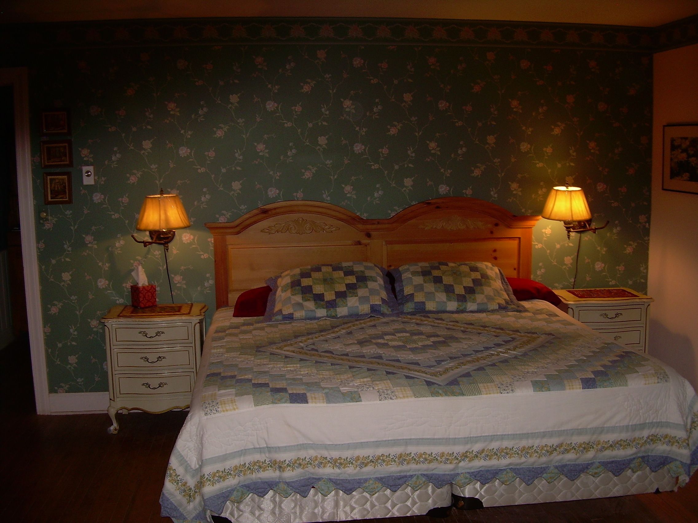 A bedroom with floral wallpaper, a wooden headboard, two bedside tables with lamps, and a quilted bedspread.