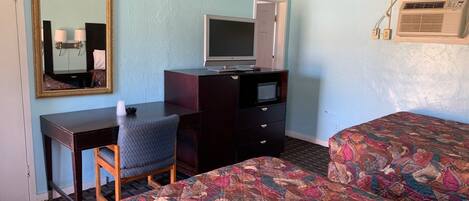 A hotel room with a bed, a desk, a chair, a television, a dresser, and an air conditioning unit.