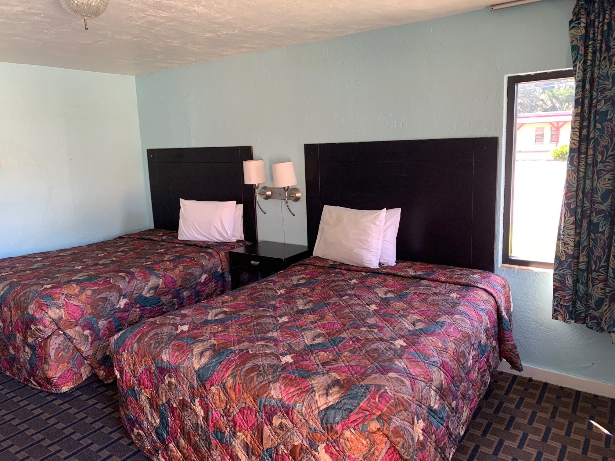 A hotel room with two beds, each with a patterned bedspread, a nightstand with two lamps, and a window with curtains.
