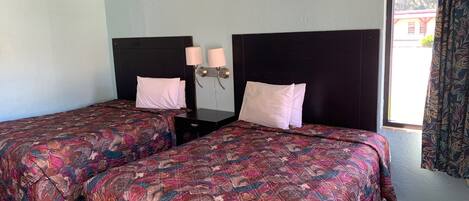 A hotel room with two beds, each with a patterned bedspread, a nightstand with two lamps, and a window with curtains.