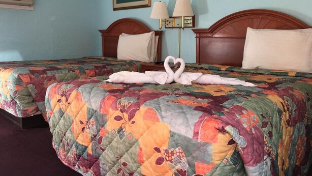Une chambre d’hôtel avec deux lits, chacun agrémenté d’une couverture colorée et d’une serviette pliée en forme de cœur. Des lampes murales et un tableau encadré sont fixés au mur au-dessus des lits.