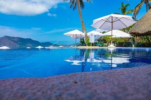 Une piscine d’eau cristalline, bordée de parasols et de palmiers, et entourée de montagnes.