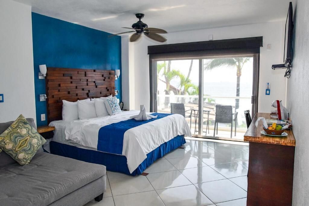 A hotel room with a bed, a gray sofa, a balcony with a view of the ocean, and a ceiling fan.
