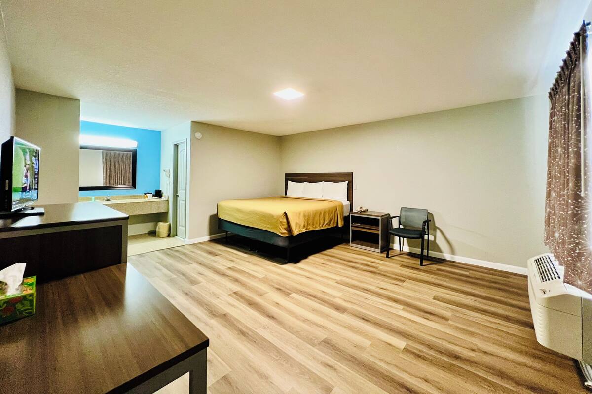 Habitación de hotel con cama, mesita de noche, silla, escritorio, televisor y baño a la vista al fondo.