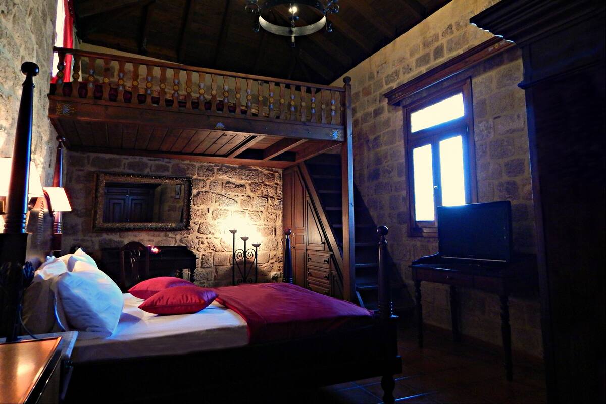 A rustic room with a wooden bed, a stone wall, a fireplace, and a wooden staircase.