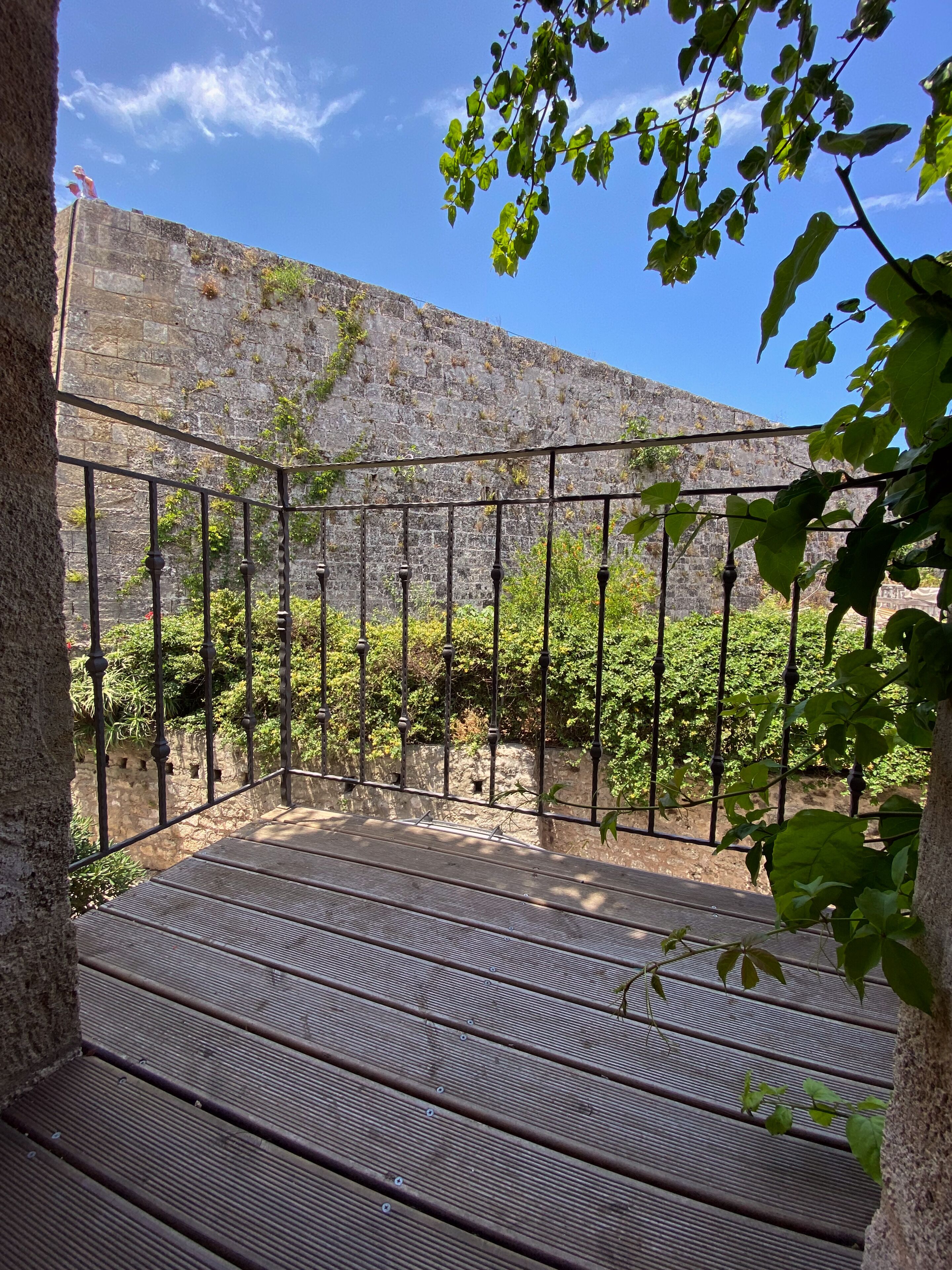 Una terraza de madera con barandas metálicas, rodeada de follaje y un muro de piedra.