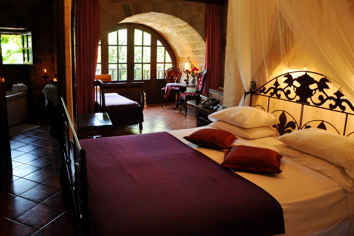 A bedroom with a large bed, a chair, a table with a vase, and a window with a stone arch.