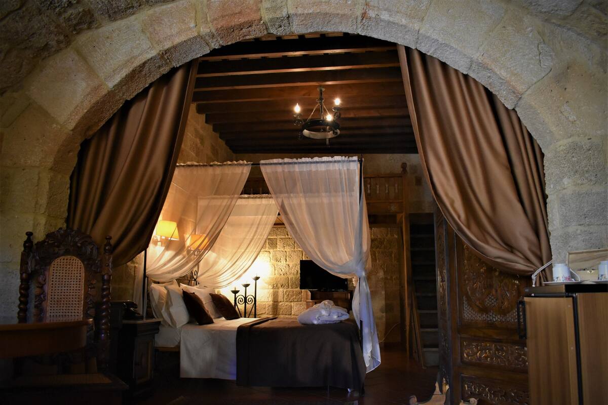A room with a stone archway, a bed with a canopy, a wooden chair, a nightstand, a lamp, and a television.
