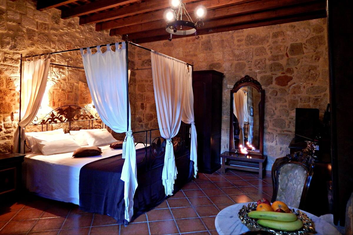 A stone-walled bedroom with a four-poster bed, a mirror, and a fruit bowl on the floor.