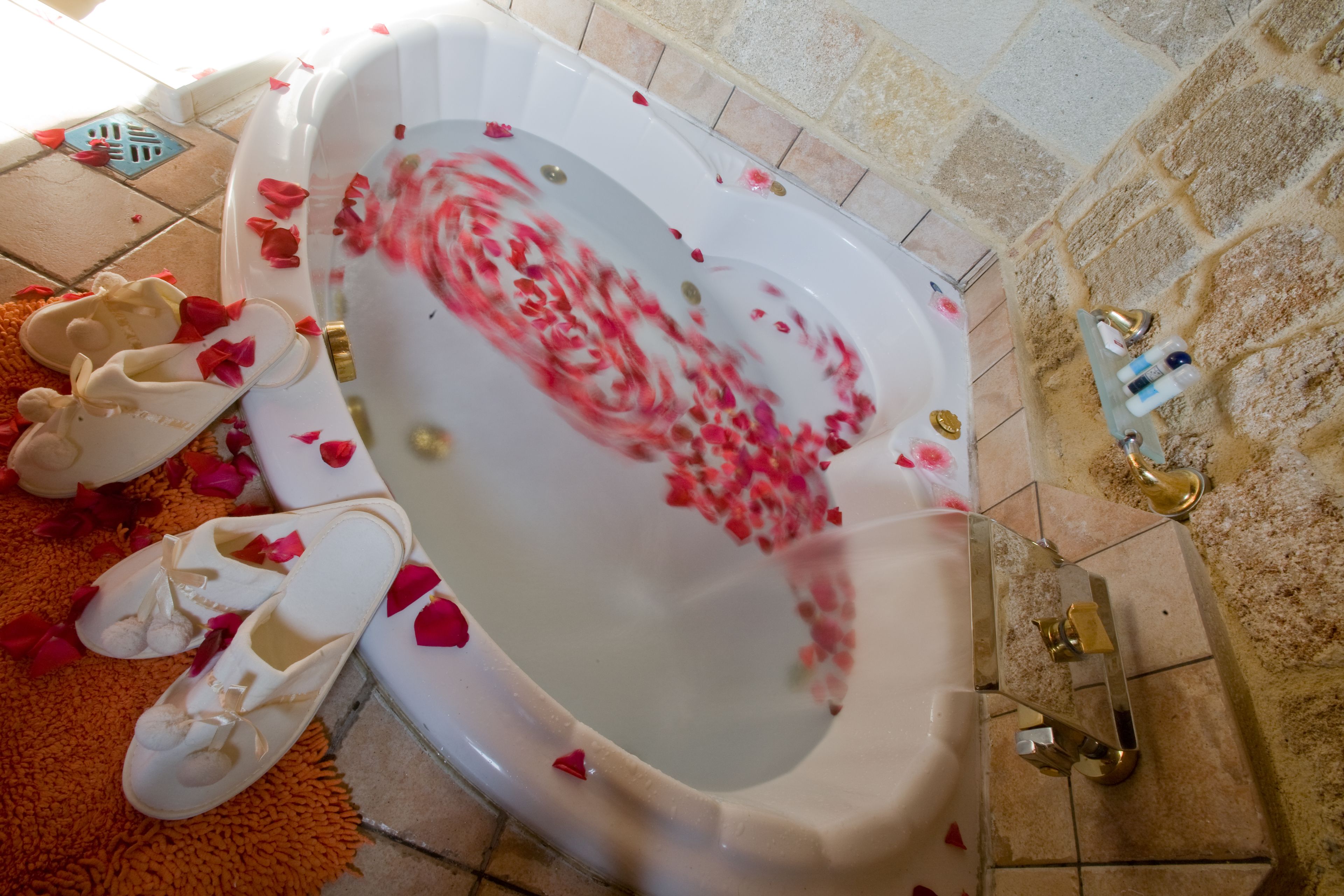 A bathtub filled with water and rose petals, with a pair of white slippers beside it.