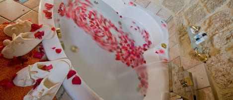 A bathtub filled with water and rose petals, with a pair of white slippers beside it.
