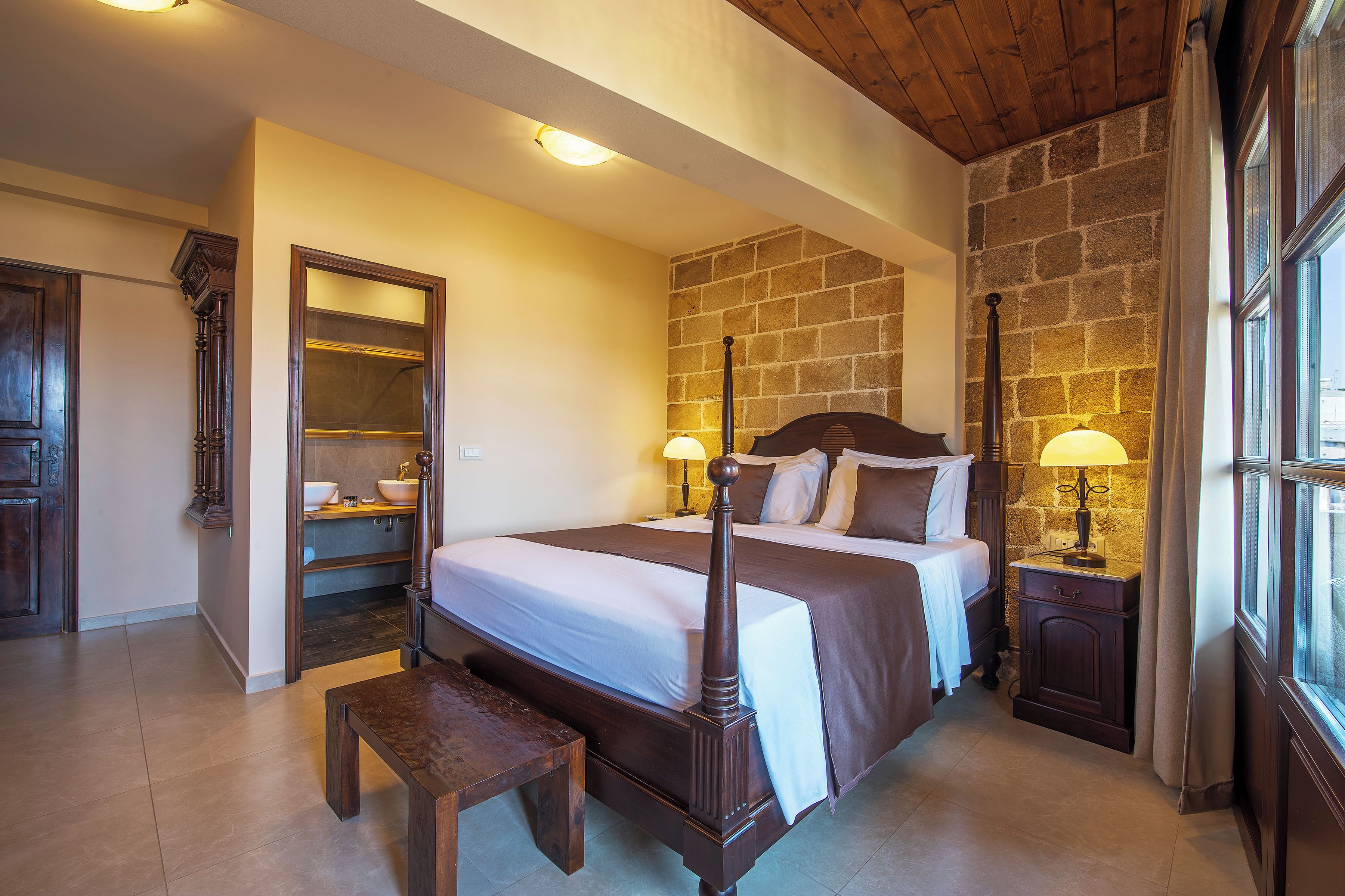 A bedroom with a four-poster bed, a wooden bench, a nightstand with a lamp, and a stone wall.