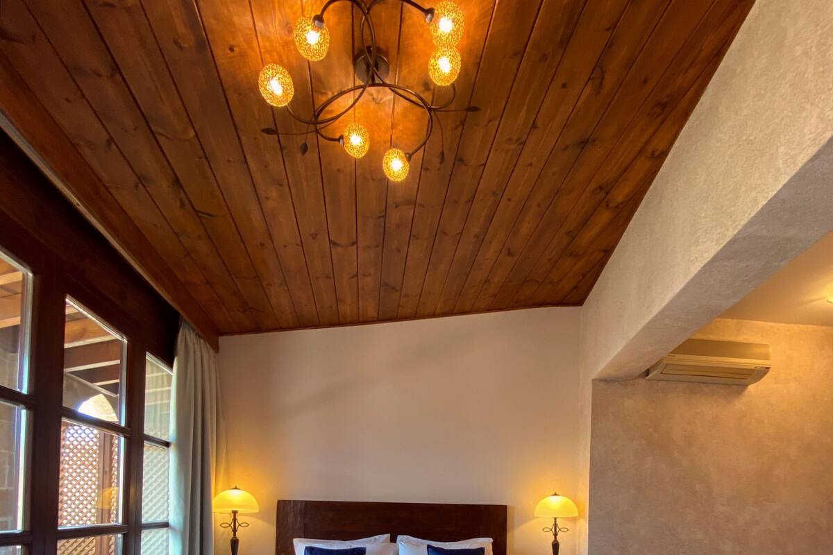 A bedroom with a wooden ceiling, a large bed with white and blue bedding, two bedside lamps, and a window with curtains.