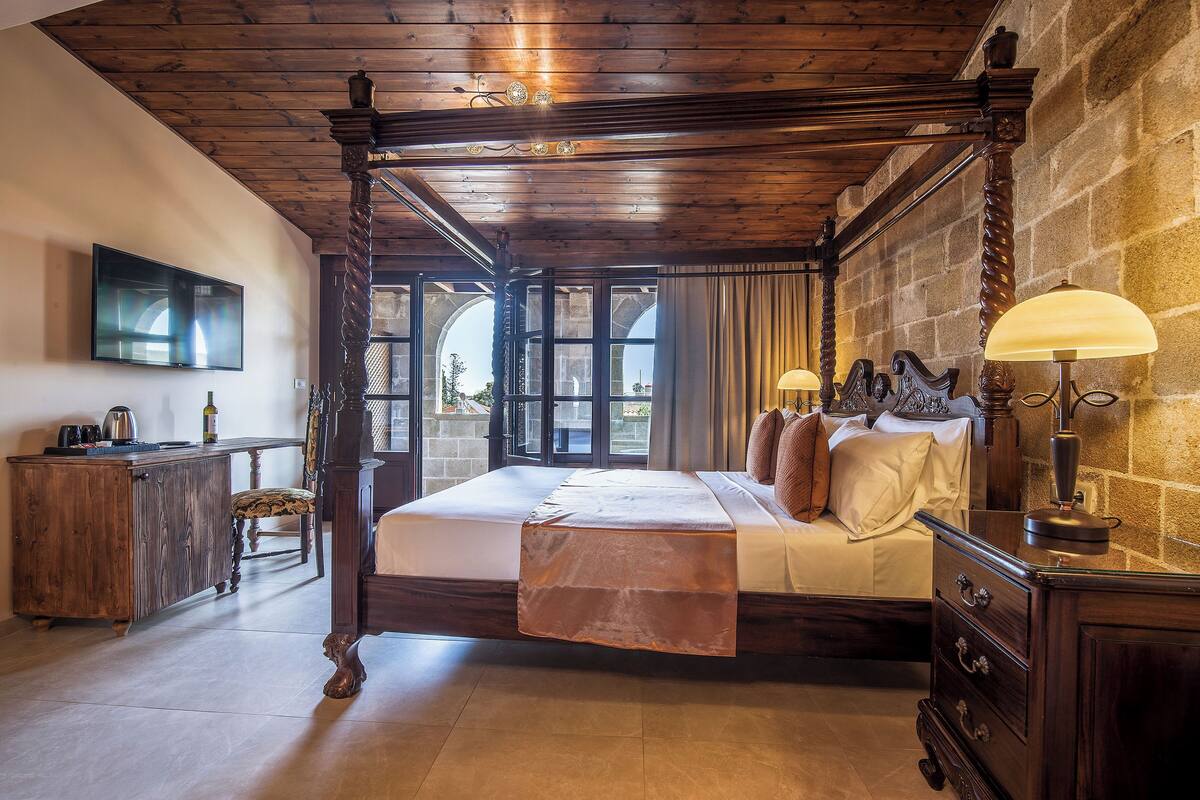 A four-poster bed with a wooden headboard, a bedside table with a lamp, and a view of the outdoors through a large window.