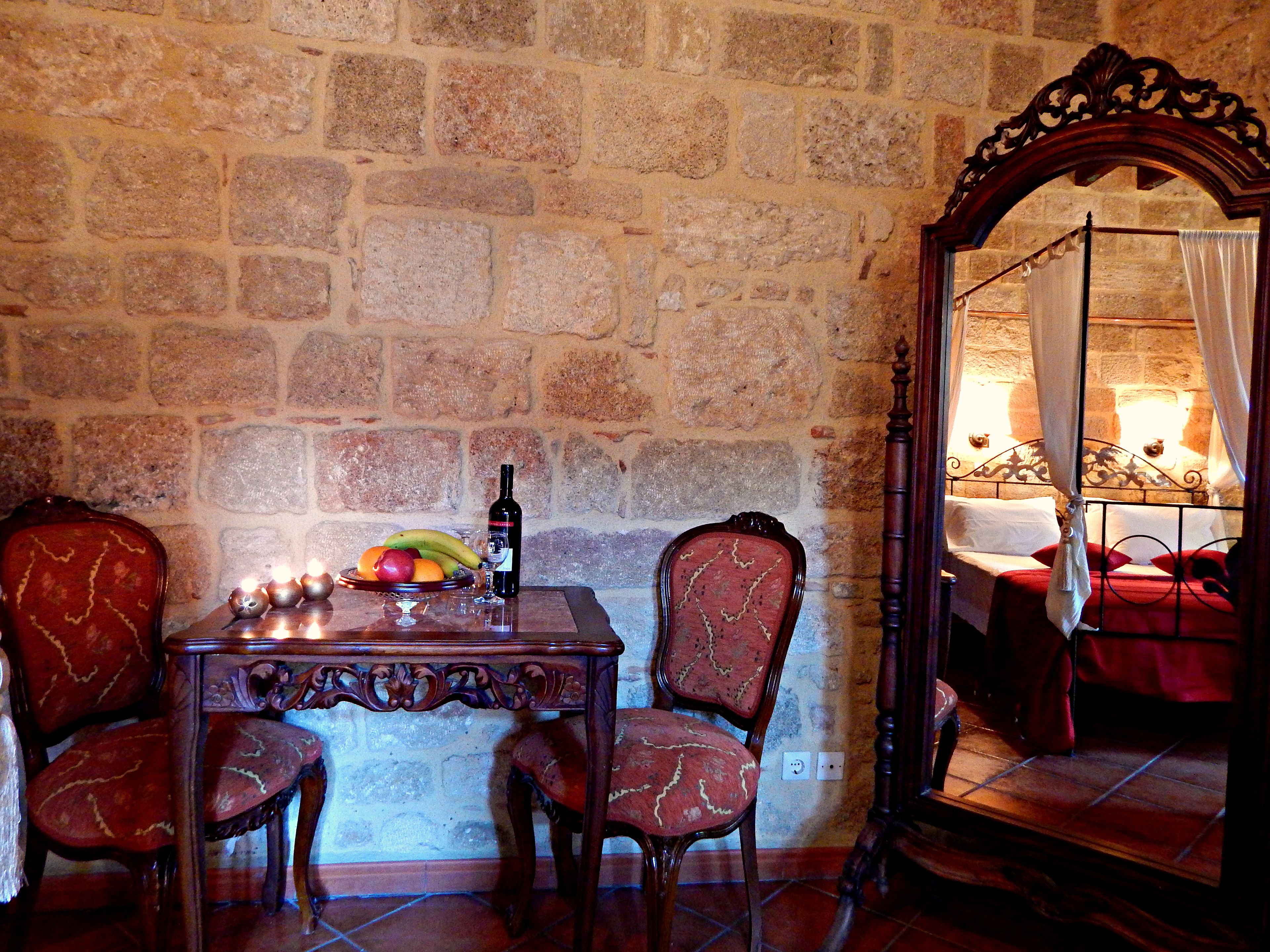 A stone-walled room with a bed, a table with fruit and a bottle, and two chairs.