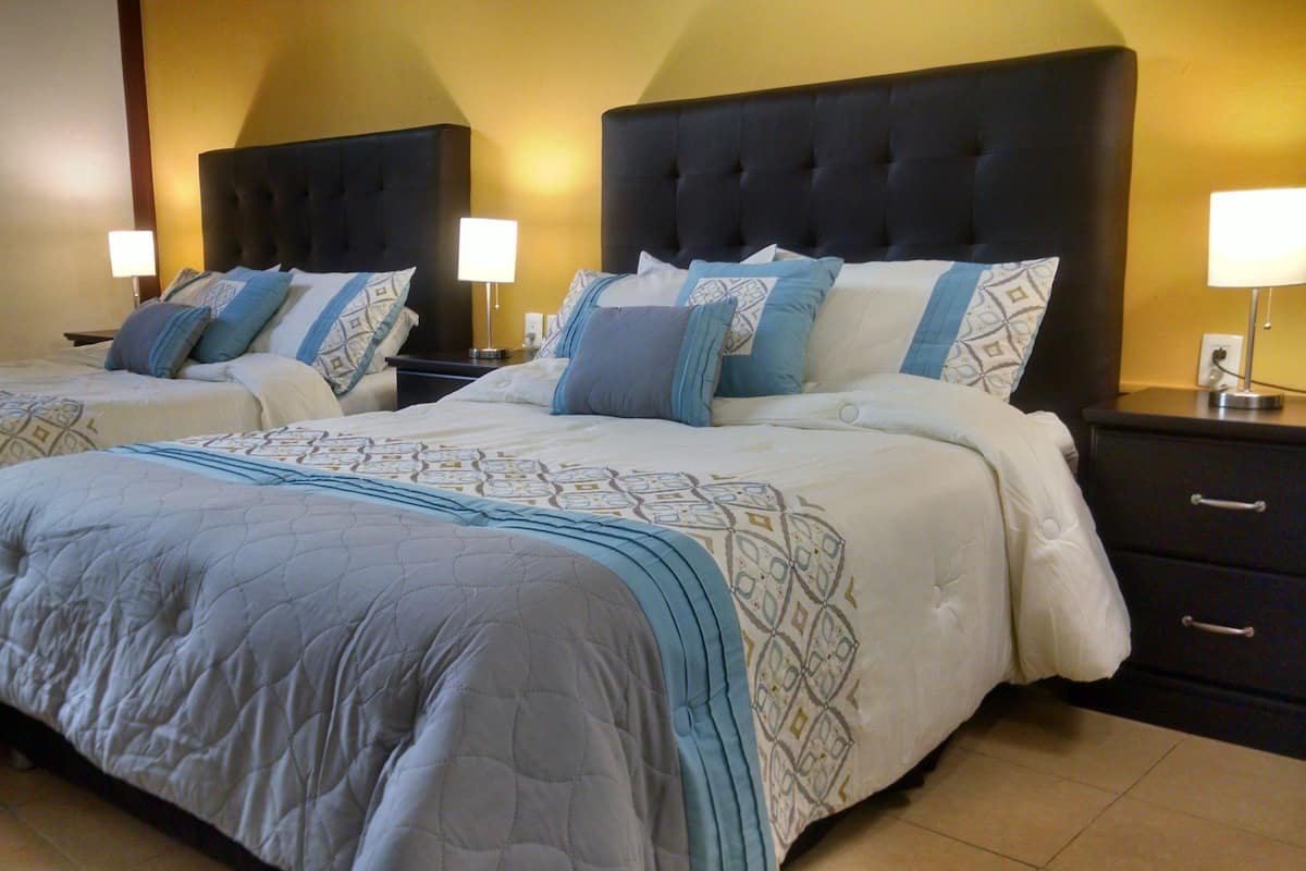 A hotel room with two queen beds, each with a headboard, bedside tables, lamps, and a nightstand.