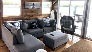 A modern living room with a grey sectional sofa, a matching ottoman, and a wicker chair. A painting of a lighthouse is on the wall, and there is a large window providing a view of the outdoors. - Mangrove Marina and Resort Aqualodge Houseboats (Tavernier)