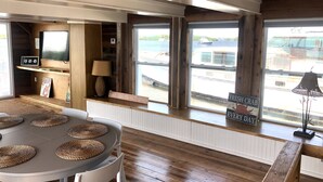 A cozy room with a wooden floor, a round table with woven placemats, a window seat with a view of the water, and a sign reading "SANDY TOES AND SALTY KISSES". - Mangrove Marina and Resort Aqualodge Houseboats (Tavernier)