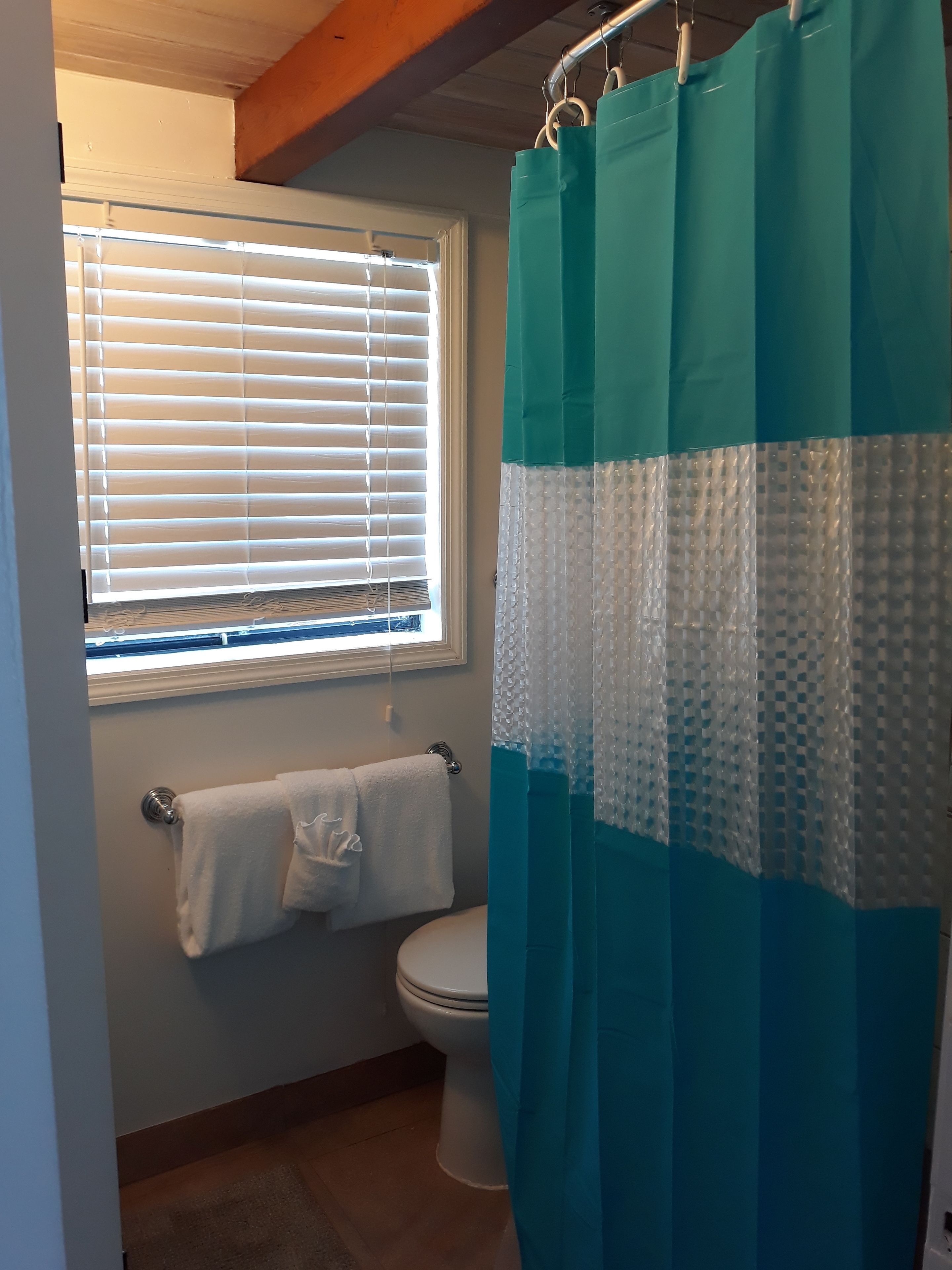 A bathroom with a toilet, a towel rack with a white towel, a window with blinds, and a shower curtain.