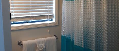 A bathroom with a toilet, a towel rack with a white towel, a window with blinds, and a shower curtain.