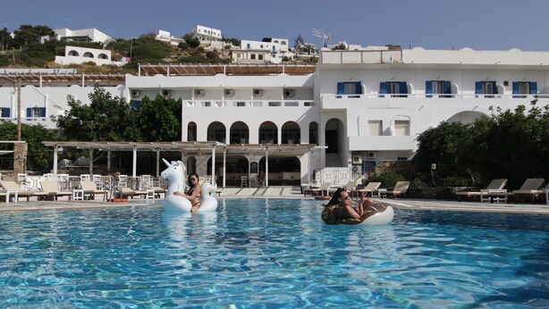 Una piscina con sillones de descanso y sombrillas, rodeada de edificios blancos con detalles azules y ventanas arqueadas.