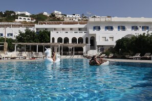 A swimming pool with lounge chairs and umbrellas, surrounded by white buildings with blue accents and arched windows. - Armadoros Hotel Ios Backpackers (Ios)