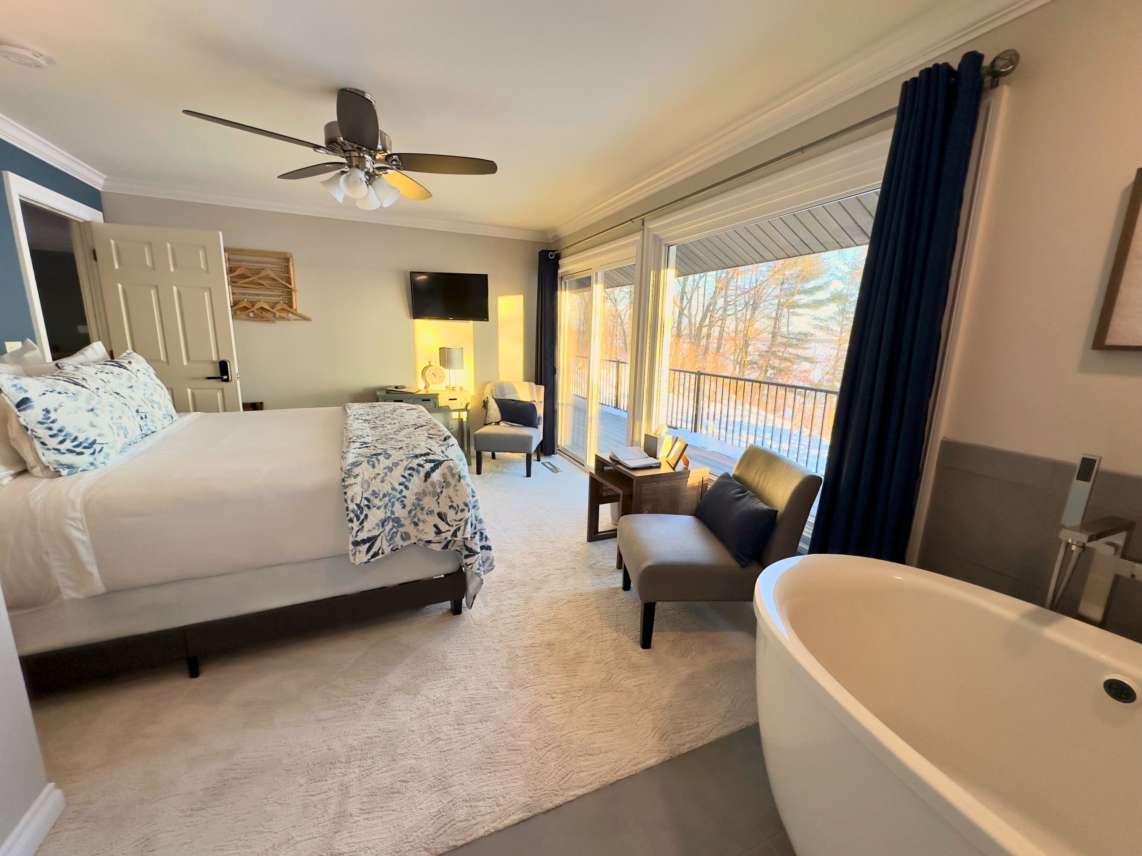 A hotel room with a bed, a sitting area with a chair and a table, a large bathtub, and a ceiling fan.