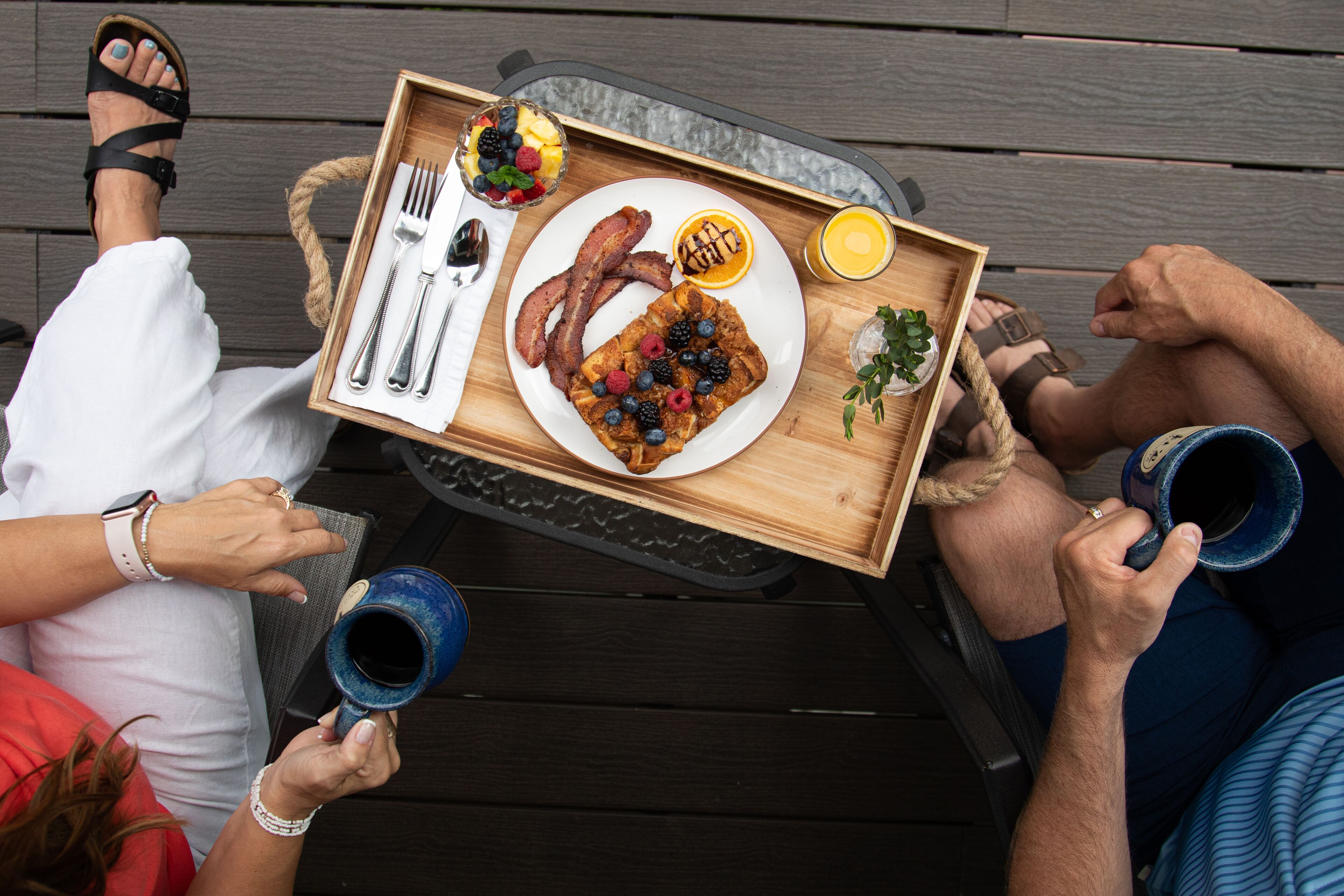 Una bandeja de madera con alimentos para el desayuno, como panqueques, tocino y fruta, sostenida por personas sentadas en una terraza de madera.