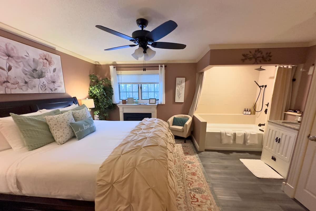 A bedroom with a bed, a chair, a desk, a window, a ceiling fan, a shower, and a bathroom.