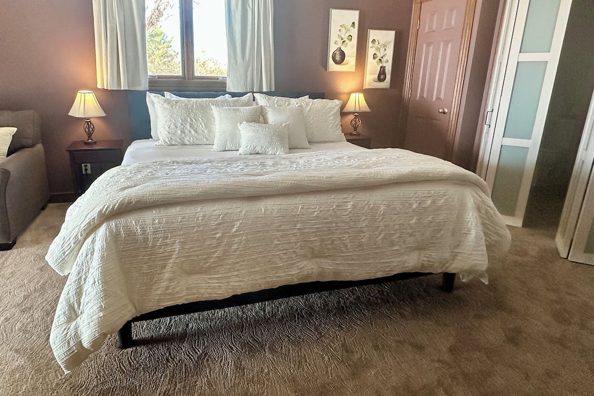 A neatly made bed with a white bedspread and several pillows, a nightstand with a lamp, and two framed pictures on the wall.