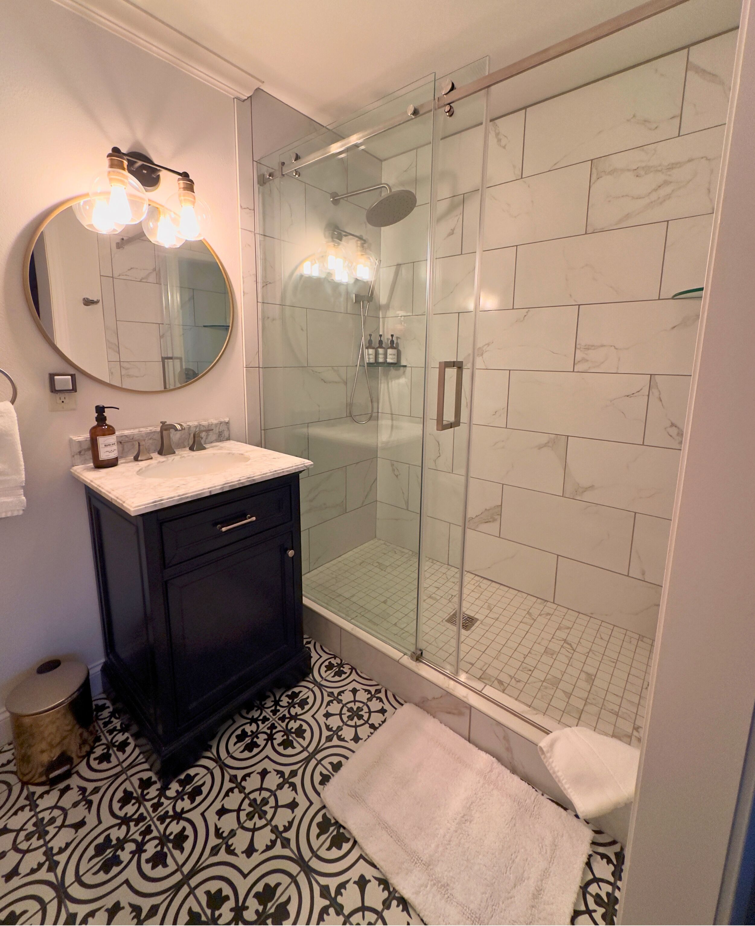 A bathroom with a glass-enclosed shower, a round mirror, and a dark cabinet.