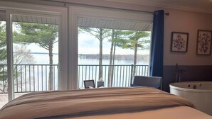 A bedroom with a bed, a chair, a bathtub, and a balcony with a view of trees and water. - Inn On Lake Wissota (Chippewa Falls)