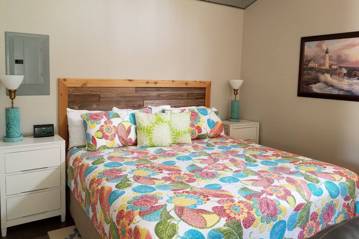 A bed with a colorful floral bedspread, wooden headboard, two bedside tables with lamps, and a painting of a lighthouse on the wall.