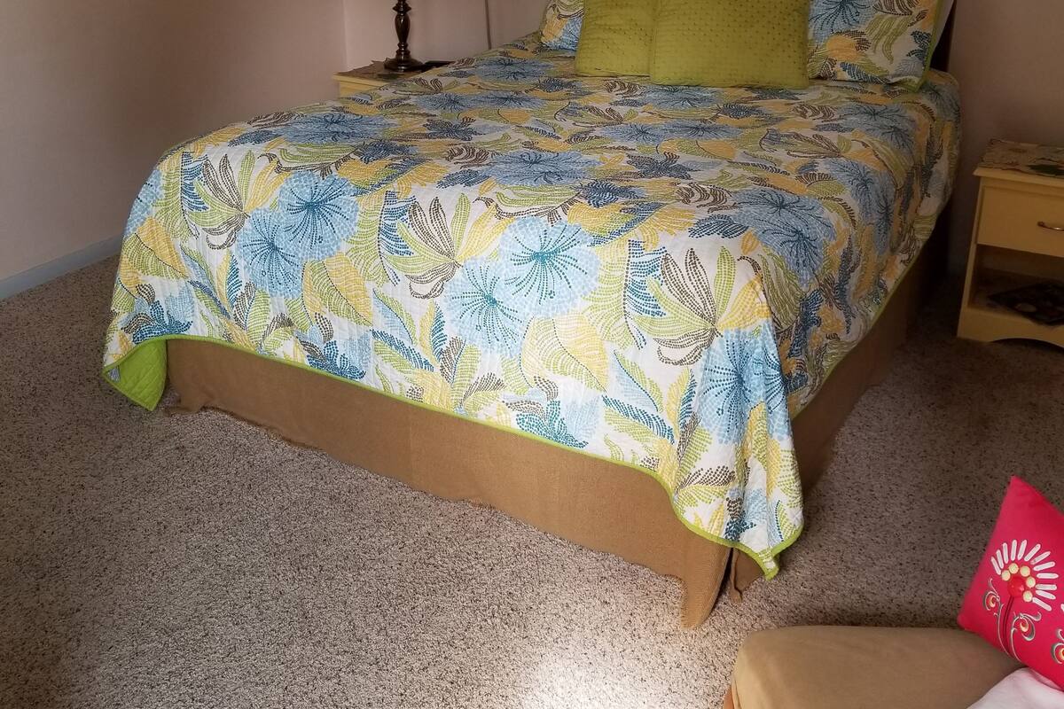 A bedroom with a floral bedspread, a window with blinds, a wall-mounted air conditioner, and a framed picture.